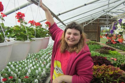 A young woman with sandy blonde hair smiles at the camera while reaching up to grab a hanging planter full of geraniums in a greenhouse